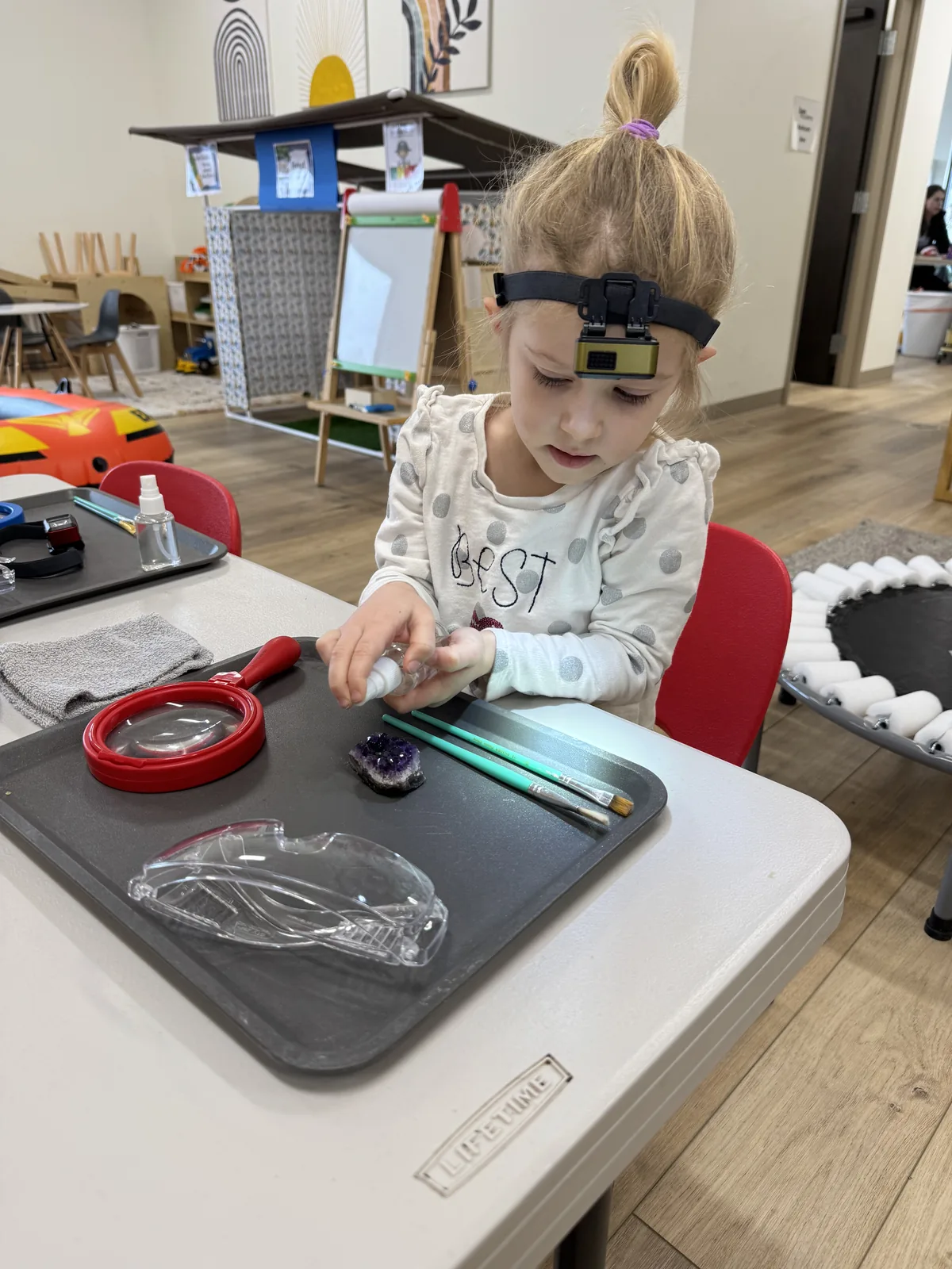 Young girl with headlamp exploring gems and minerals at a science discovery table during Discovery Station at Spark Academy